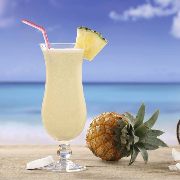 A tropical drink with pineapple and a red straw sits on a beachside table next to a whole pineapple.