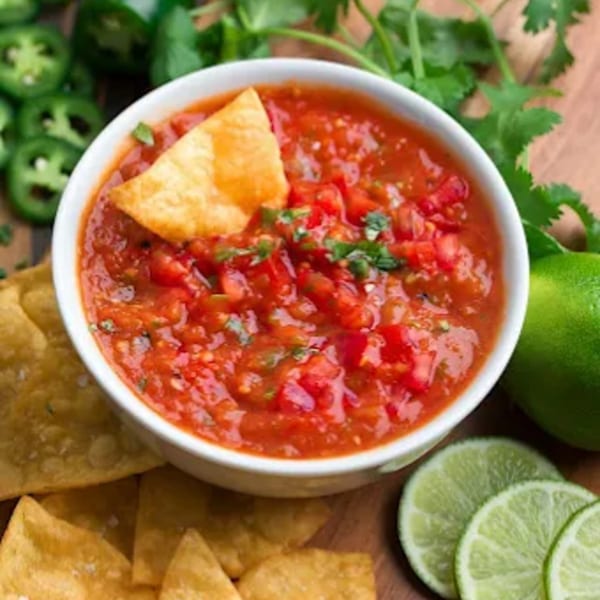 A bowl of Don Jose's fresh salsa with a tortilla chip, surrounded by limes and jalapeños.
